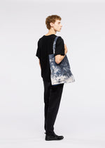 A young person with curly hair stands sideways in black attire, holding the GOOD GOODS ISSEY MIYAKE IROGAMI SALT DYE tote bag against a plain white background.