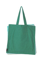 A large green IROGAMI SALT DYE tote bag by GOOD GOODS ISSEY MIYAKE, with long handles and made using the salt-dyeing technique, is shown against a white background with light blue and white bags partially visible behind it.