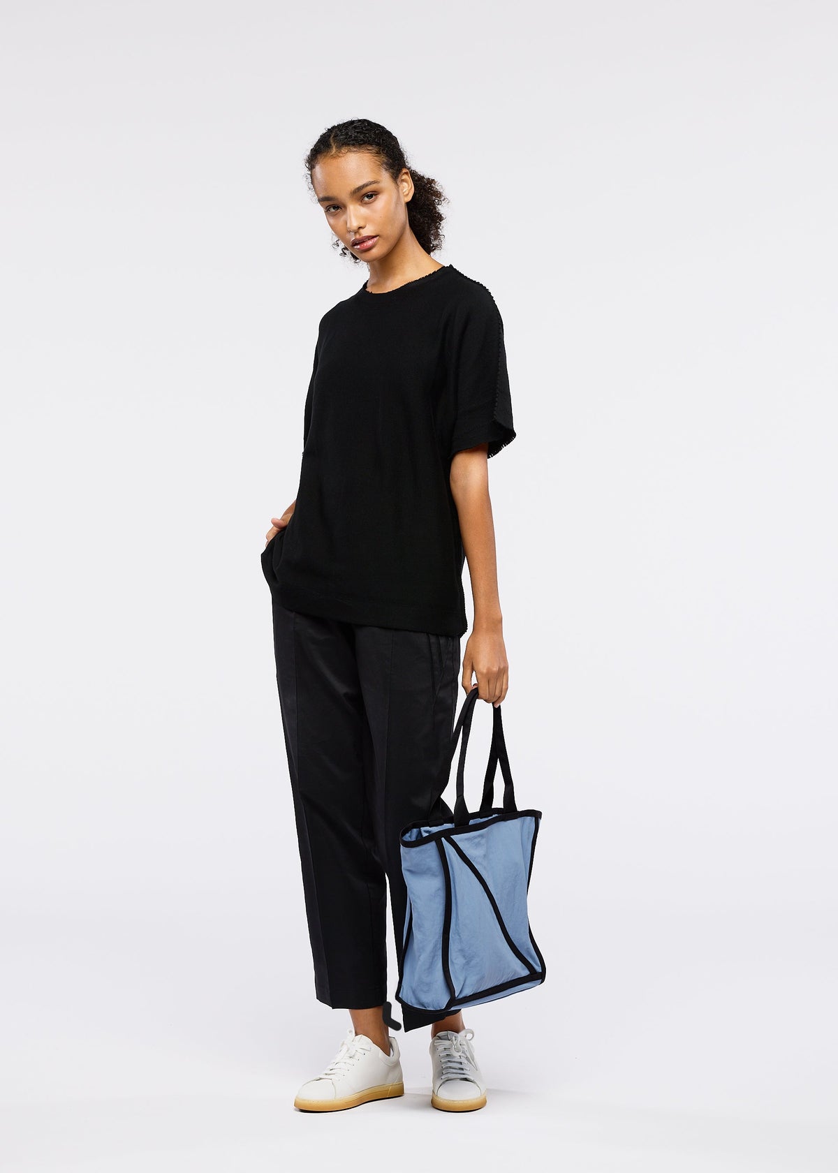 Against a plain background, a person wears a loose black t-shirt, black pants, and white sneakers, holding the GOOD GOODS ISSEY MIYAKE WAKU light blue tote bag with wide gussets in one hand and keeping their other hand in their pocket. view 6 of 7