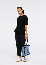 Against a plain background, a person wears a loose black t-shirt, black pants, and white sneakers, holding the GOOD GOODS ISSEY MIYAKE WAKU light blue tote bag with wide gussets in one hand and keeping their other hand in their pocket.