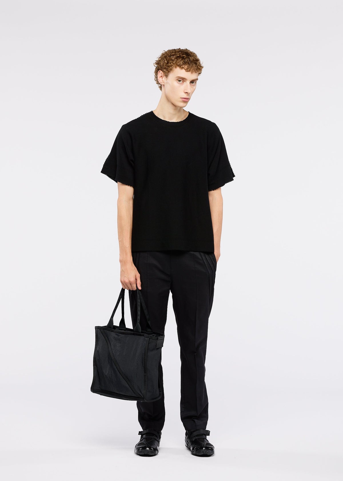 A person with short curly hair stands against a white background, wearing black clothing and holding the WAKU black tote bag with wide gussets by GOOD GOODS ISSEY MIYAKE. view 6 of 6