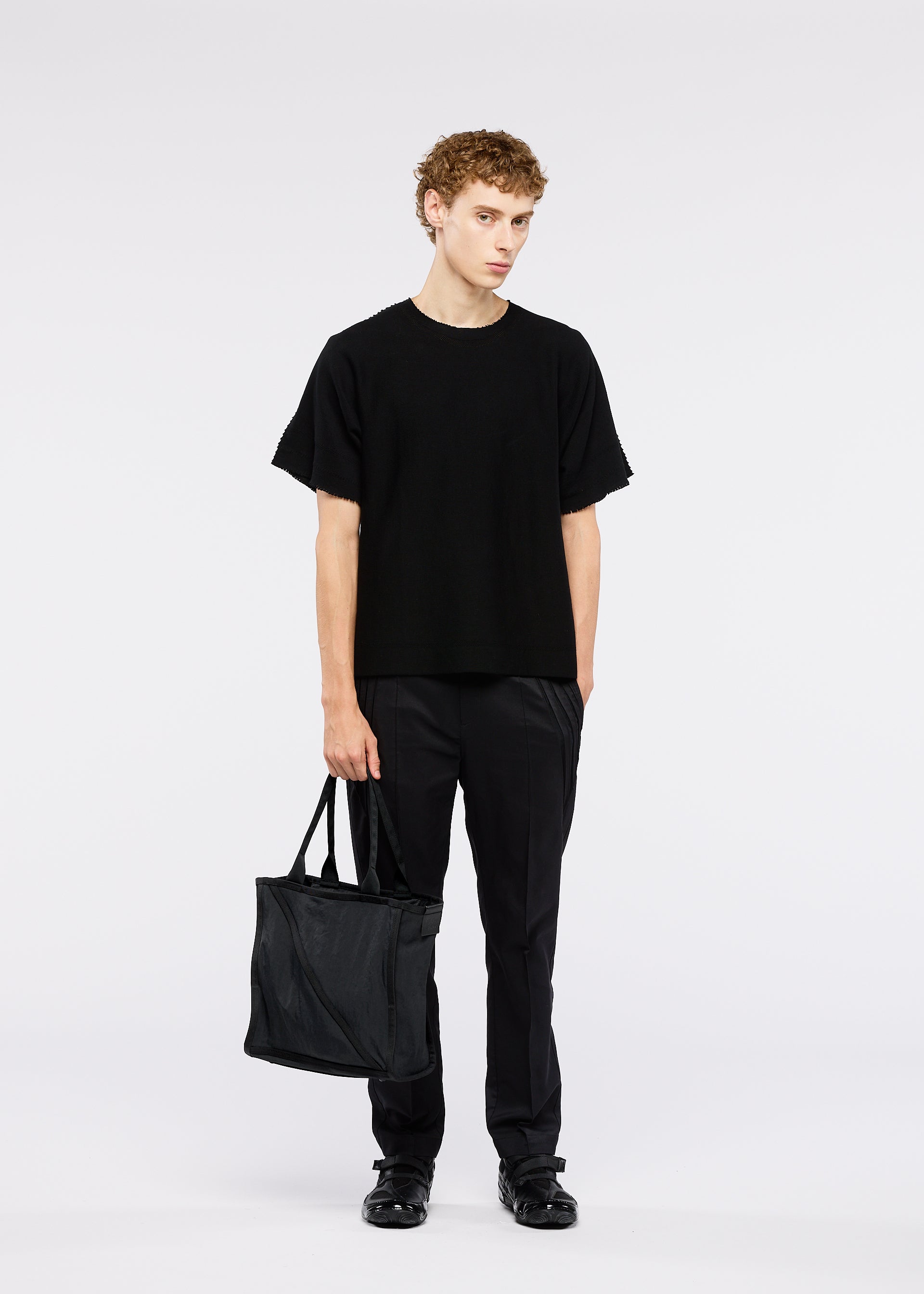 Zoomed view A person with short curly hair stands against a white background, wearing black clothing and holding the WAKU black tote bag with wide gussets by GOOD GOODS ISSEY MIYAKE.
