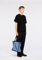 A young person stands against a white background holding the WAKU tote bag by GOOD GOODS ISSEY MIYAKE—light blue with wide gussets and black trim—paired with a black t-shirt, pants, and shoes.