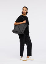 A person stands against a plain white background, wearing black attire and white sneakers, carrying the WAKU tote bag by GOOD GOODS ISSEY MIYAKE with wide gussets, looking off to the side with a neutral expression.