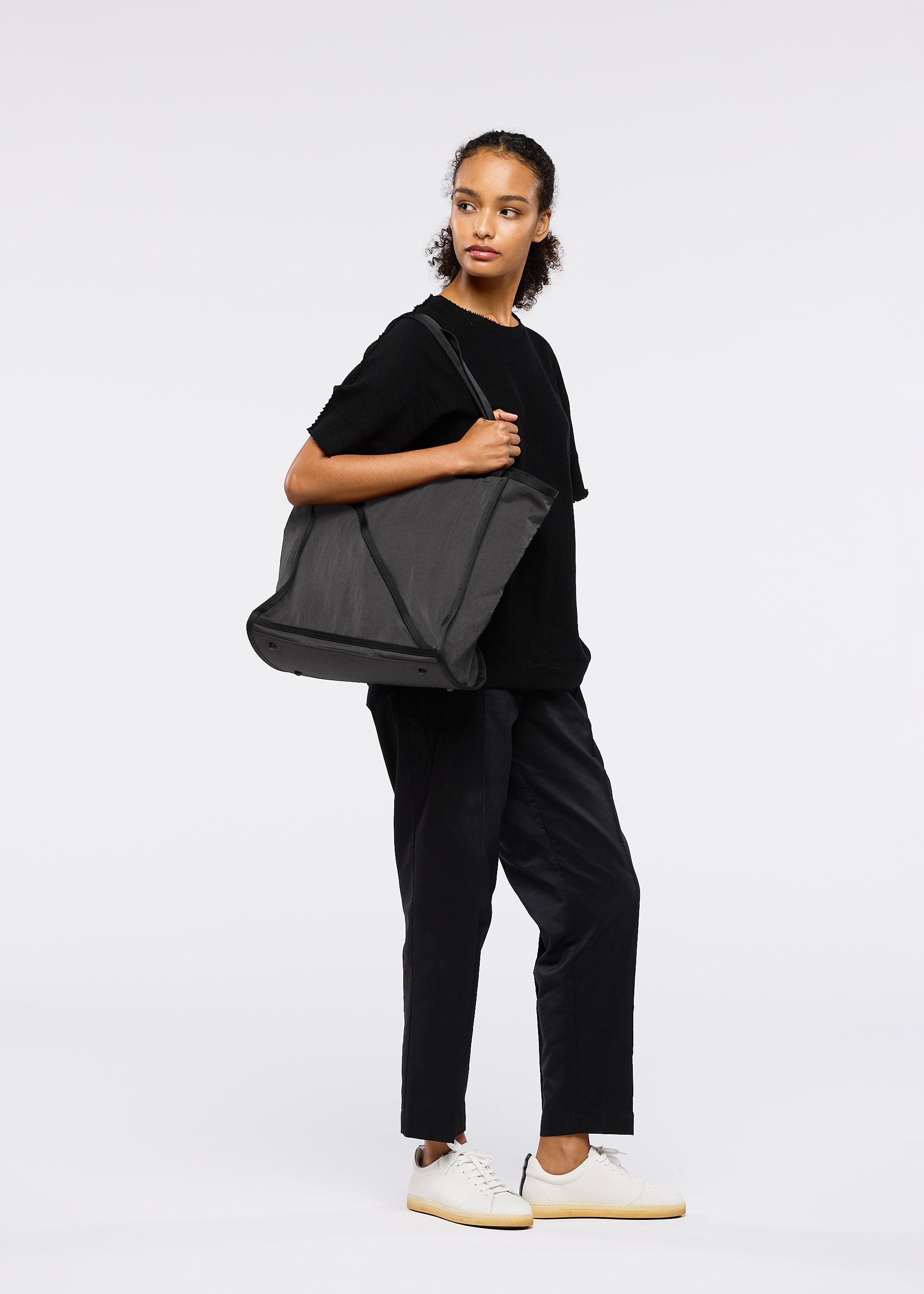 Zoomed view A person stands against a plain white background, wearing black attire and white sneakers, carrying the WAKU tote bag by GOOD GOODS ISSEY MIYAKE with wide gussets, looking off to the side with a neutral expression.