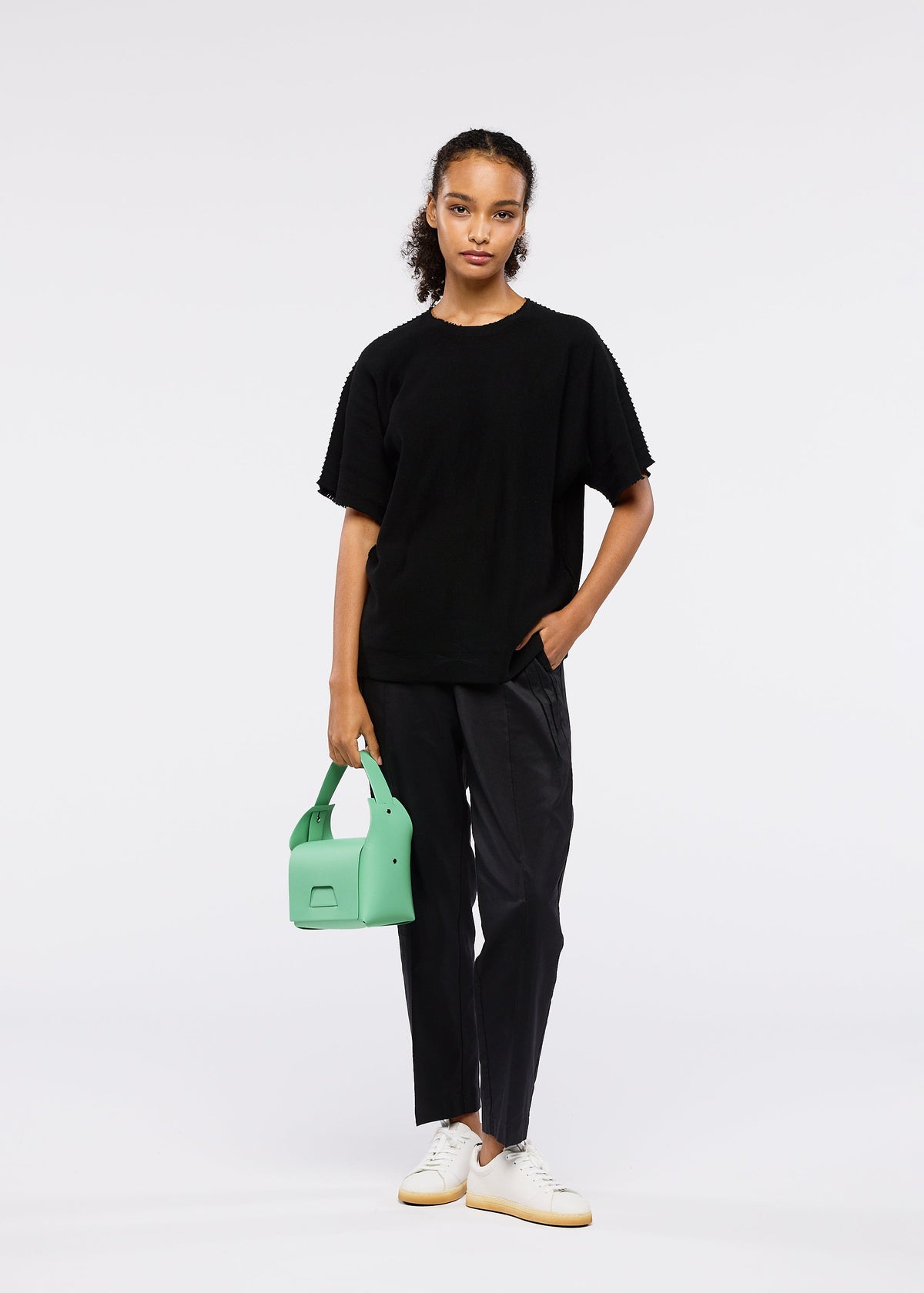 A woman stands against a plain white background wearing black pants, a loose black t-shirt, and white sneakers. She holds the light green BOX bag from GOOD GOODS ISSEY MIYAKE with her hair pulled back. view 8 of 10