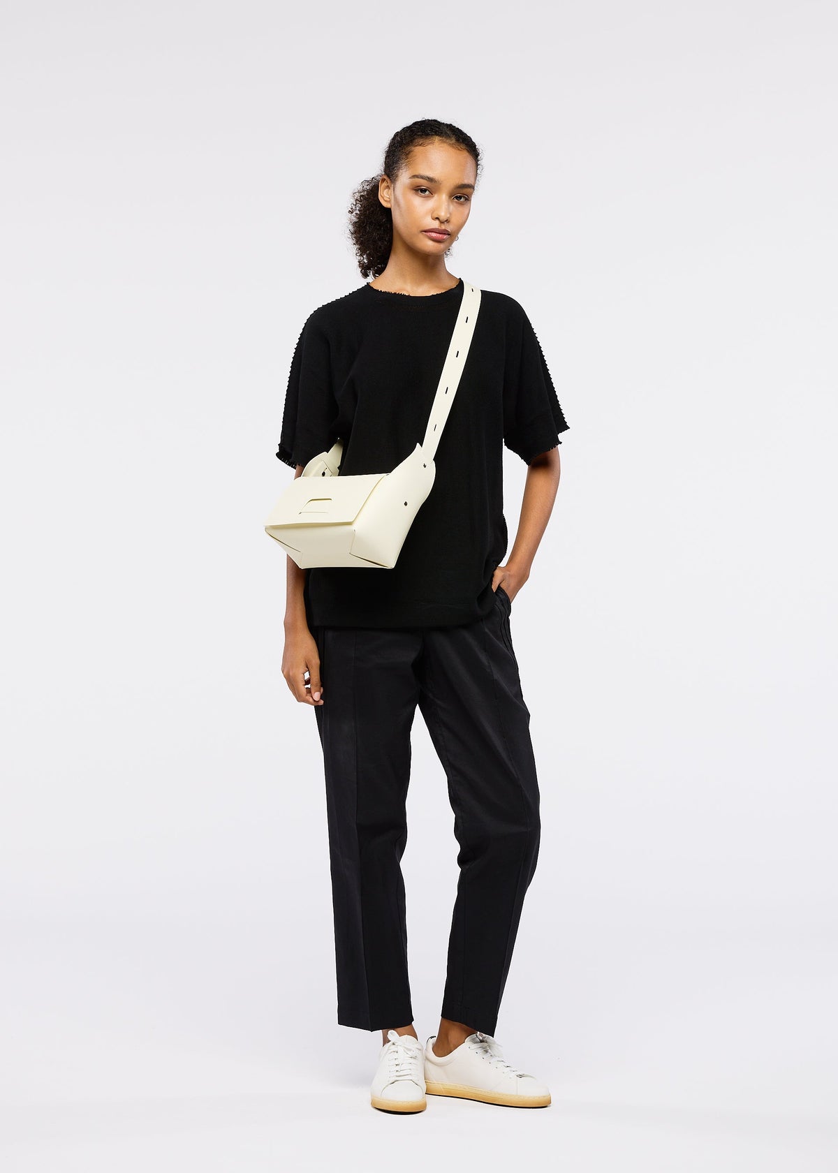 A woman with a neutral expression stands against a white background wearing black trousers, a loose black T-shirt, white sneakers, and the cream BOX bag by GOOD GOODS ISSEY MIYAKE, her left hand casually in her pocket. view 10 of 11