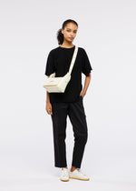 A woman with a neutral expression stands against a white background wearing black trousers, a loose black T-shirt, white sneakers, and the cream BOX bag by GOOD GOODS ISSEY MIYAKE, her left hand casually in her pocket.