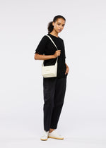 A woman poses against a plain background, wearing black pants, a black tee, white sneakers, and carries the GOOD GOODS ISSEY MIYAKE BOX cream convertible bag over her shoulder. One hand rests in her pocket as she stands confidently.