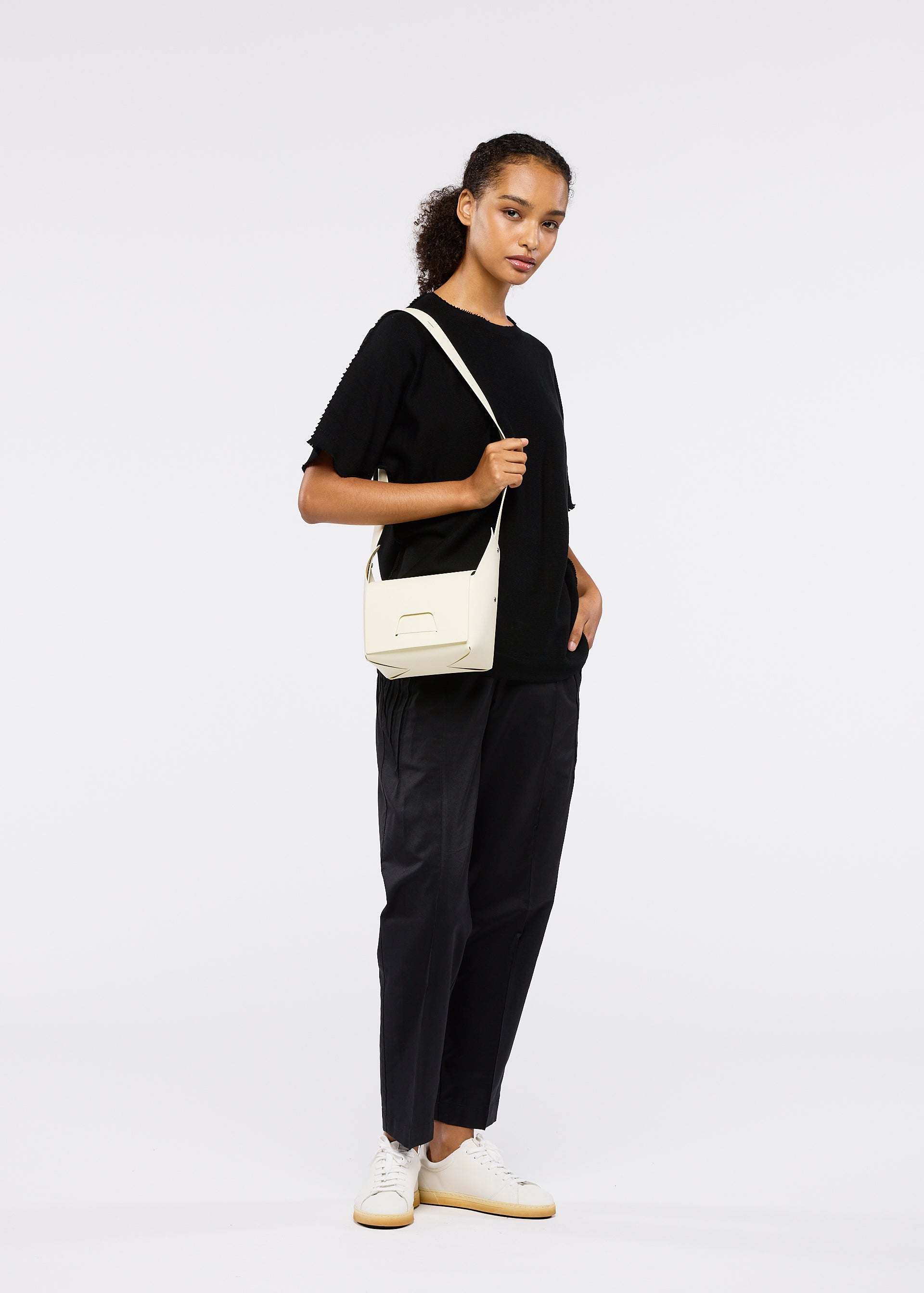Zoomed view A woman stands against a white background, wearing black attire and white sneakers. She carries the BOX bag from GOOD GOODS ISSEY MIYAKE on her shoulder, hair tied back, one hand in her pocket, looking at the camera.