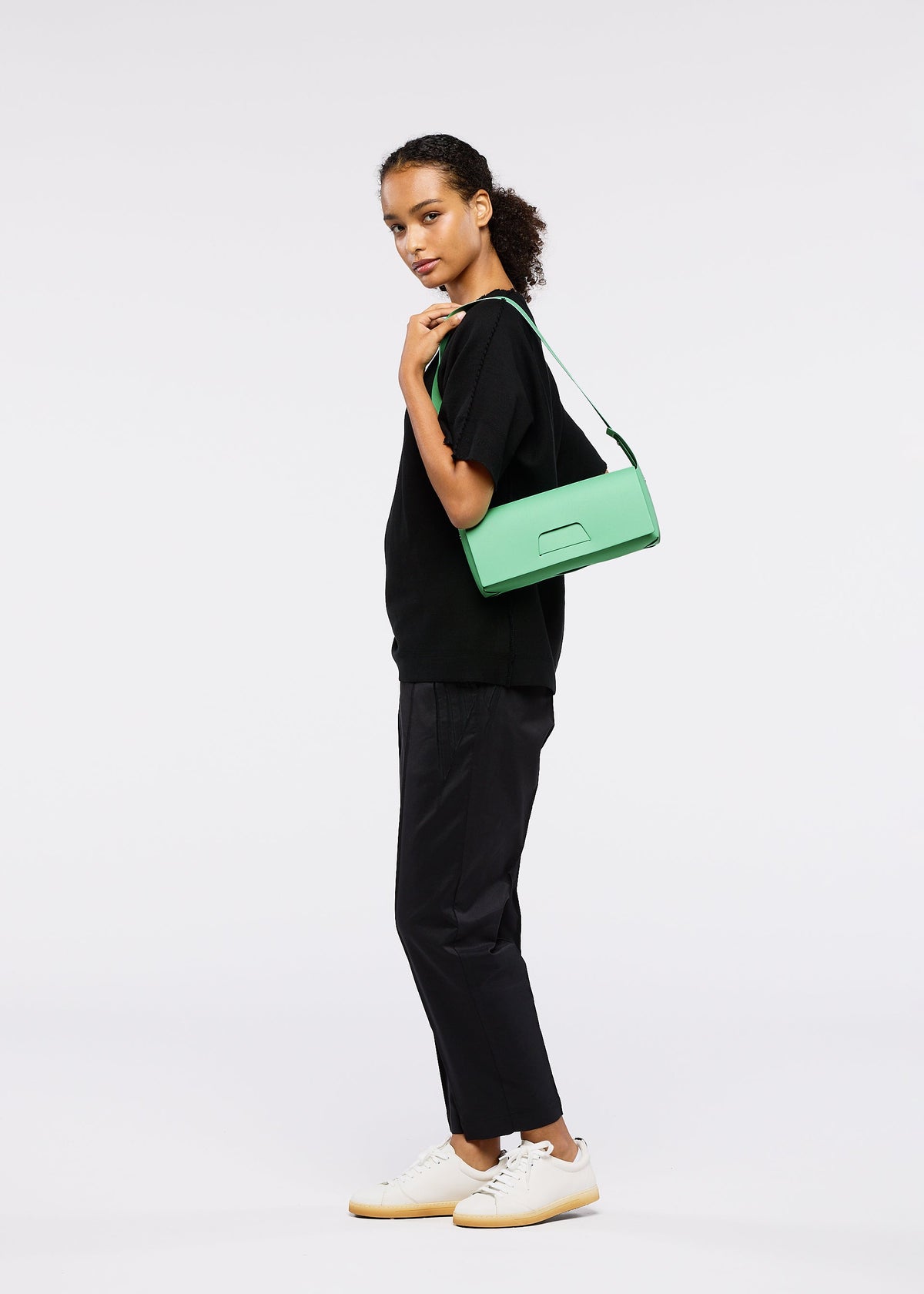 A person stands sideways against a plain background, wearing a loose black top, black pants, white sneakers, and carrying the GOOD GOODS ISSEY MIYAKE BOX 26SS new model mint green convertible shoulder bag. view 7 of 9