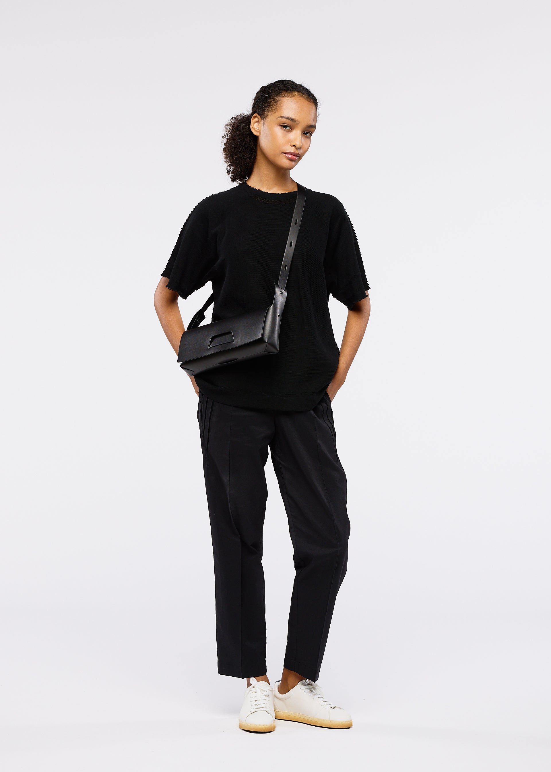 Zoomed view A person poses in a black short-sleeve top, loose black pants, white sneakers, and the GOOD GOODS ISSEY MIYAKE BOX convertible bag—26SS model—against a plain white background.