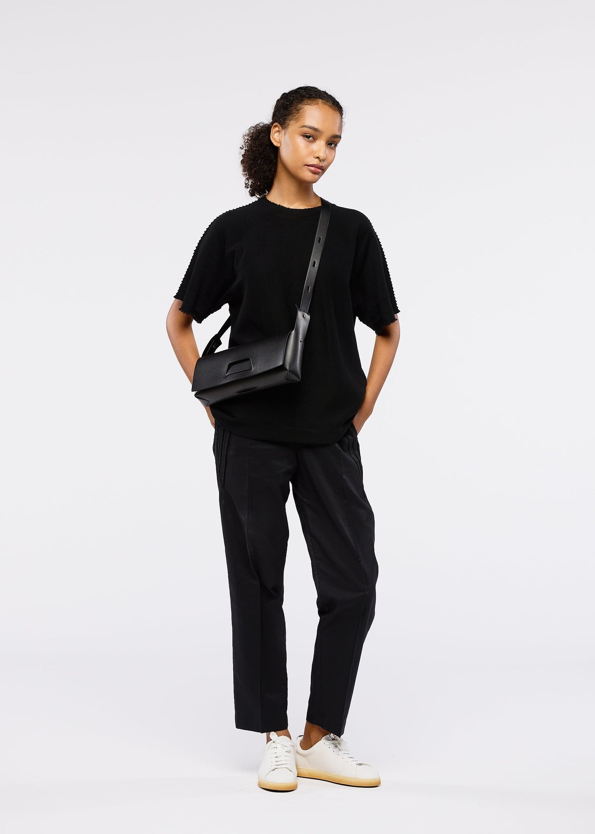 A person stands against a plain white background, wearing a black short-sleeve top, black pants, white sneakers, and the 26SS BOX convertible shoulder bag by GOOD GOODS ISSEY MIYAKE. Their curly hair is tied back in a relaxed, confident pose. view 8 of 9