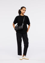 A person stands against a plain white background, wearing a black short-sleeve top, black pants, white sneakers, and the 26SS BOX convertible shoulder bag by GOOD GOODS ISSEY MIYAKE. Their curly hair is tied back in a relaxed, confident pose.
