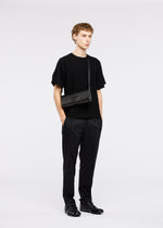 A young person with short curly hair stands facing forward, wearing black clothing and black shoes, carrying the BOX convertible shoulder bag from GOOD GOODS ISSEY MIYAKE against a plain white background.