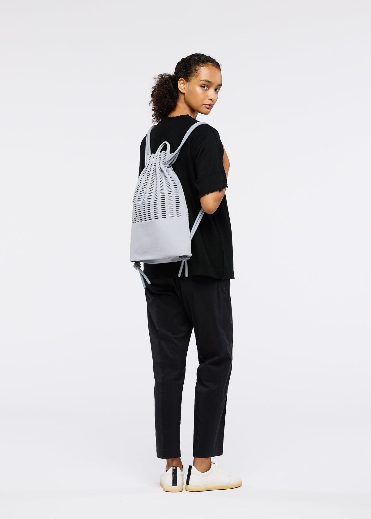 A person with curly hair, wearing a black t-shirt, black pants, and white sneakers, stands sideways glancing back while carrying the MOKKO KNAPSACK by GOOD GOODS ISSEY MIYAKE. The stylish knit bag features an adjustable shoulder strap. Plain white background. view 5 of 6