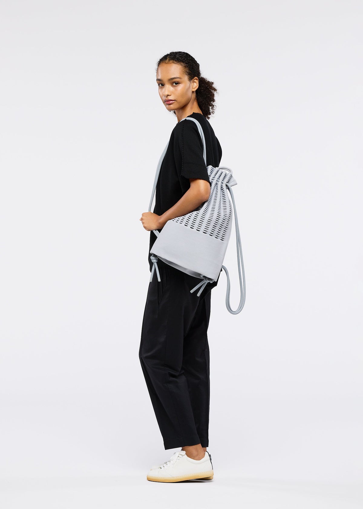 A woman with curly hair, dressed in black attire and white sneakers, stands sideways holding the MOKKO KNAPSACK by GOOD GOODS ISSEY MIYAKE against a plain white background. view 6 of 6