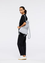 A woman with curly hair, dressed in black attire and white sneakers, stands sideways holding the MOKKO KNAPSACK by GOOD GOODS ISSEY MIYAKE against a plain white background.
