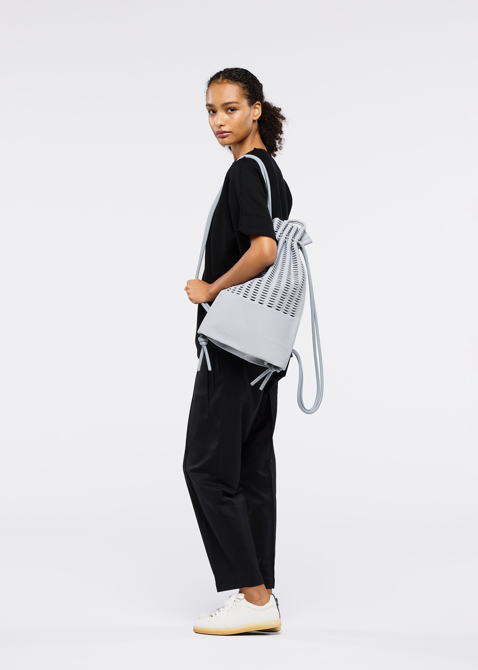 Zoomed view A woman with curly hair, dressed in black attire and white sneakers, stands sideways holding the MOKKO KNAPSACK by GOOD GOODS ISSEY MIYAKE against a plain white background.