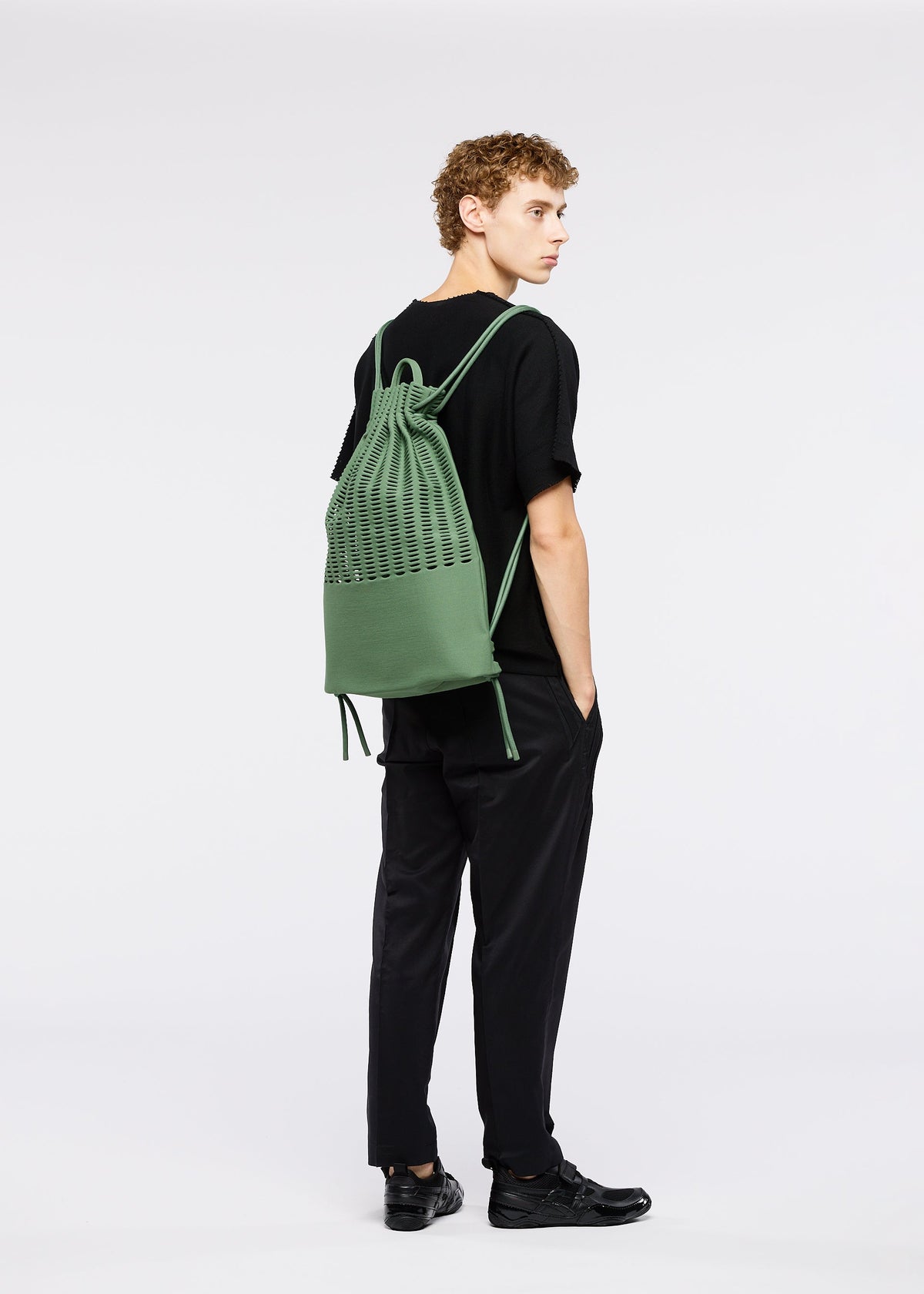 A young man with curly hair, dressed in black, stands sideways with hands in pockets, carrying the GOOD GOODS ISSEY MIYAKE MOKKO KNAPSACK—a large green knit bag with an adjustable shoulder strap—against a plain white background. view 5 of 6