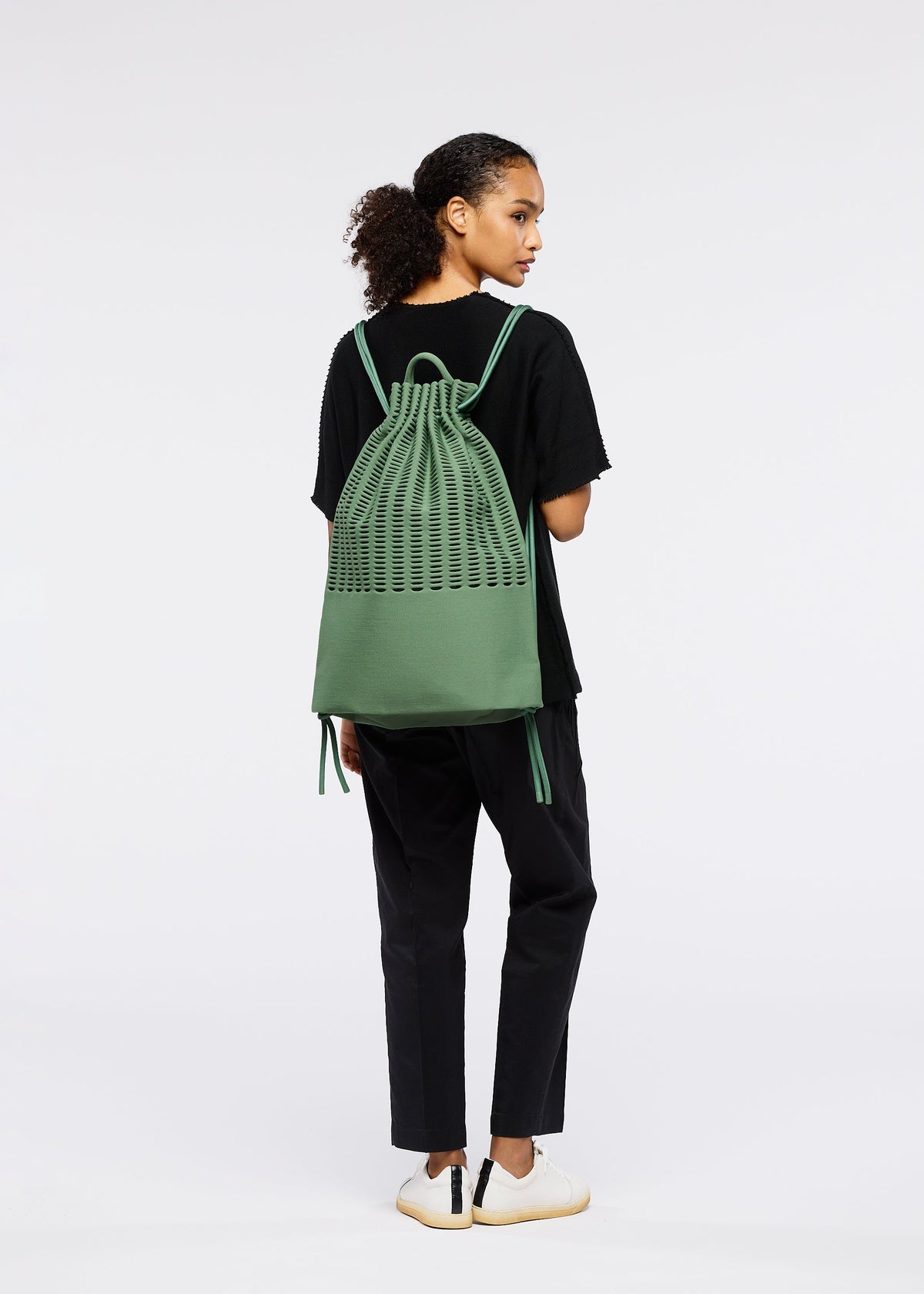 A person with curly hair in a ponytail, facing away, wears a black t-shirt, black pants, white sneakers, and carries the green MOKKO KNAPSACK by GOOD GOODS ISSEY MIYAKE with an adjustable strap against a plain white background. view 6 of 6
