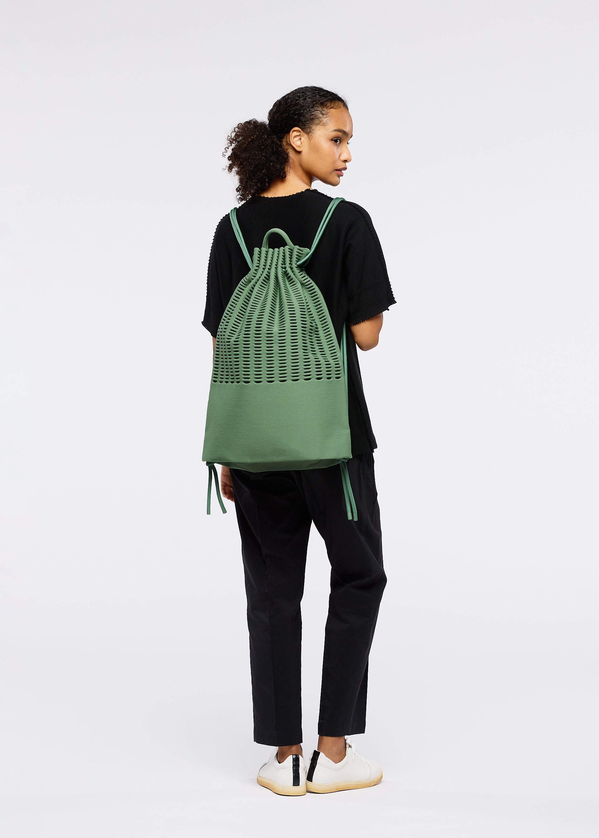 Zoomed view A person with curly hair in a ponytail, facing away, wears a black t-shirt, black pants, white sneakers, and carries the green MOKKO KNAPSACK by GOOD GOODS ISSEY MIYAKE with an adjustable strap against a plain white background.