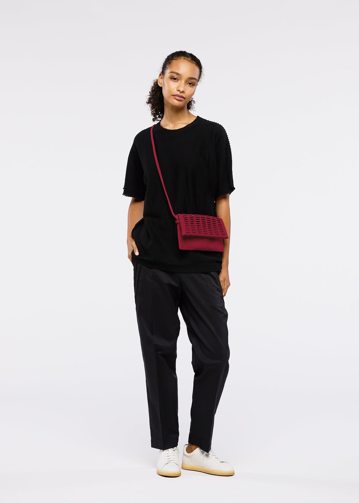 A person with curly hair pulled back poses casually against a white background, wearing a loose black t-shirt, dark pants, white sneakers, and carrying the GOOD GOODS ISSEY MIYAKE MOKKO FLAP POUCH in red. view 8 of 12