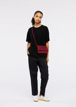 A person with curly hair pulled back poses casually against a white background, wearing a loose black t-shirt, dark pants, white sneakers, and carrying the GOOD GOODS ISSEY MIYAKE MOKKO FLAP POUCH in red.