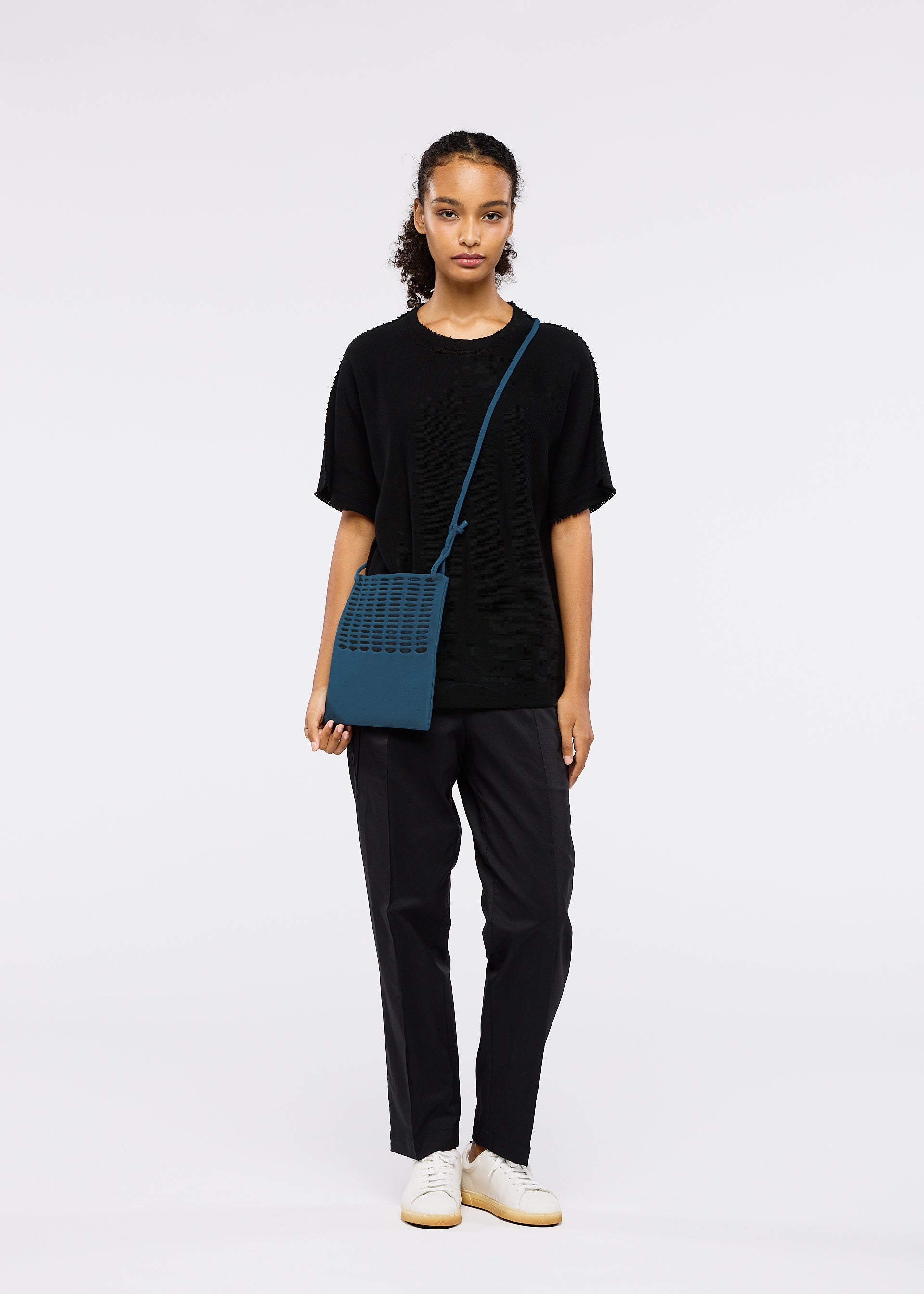 Zoomed view A woman with curly hair tied back and a neutral expression stands against a white background, wearing black attire and white sneakers, carrying the MOKKO FLAP POUCH by GOOD GOODS ISSEY MIYAKE in blue.