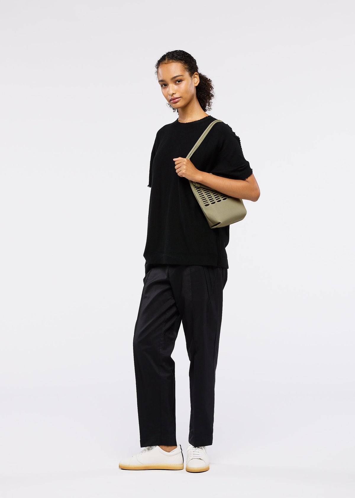 A person stands facing forward against a white background, wearing a loose black t-shirt, black pants, white sneakers, and carrying the GOOD GOODS ISSEY MIYAKE MOKKO FLAP POUCH over one shoulder. view 10 of 12