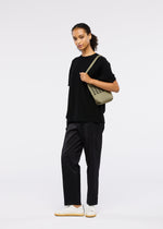 A person stands facing forward against a white background, wearing a loose black t-shirt, black pants, white sneakers, and carrying the GOOD GOODS ISSEY MIYAKE MOKKO FLAP POUCH over one shoulder.