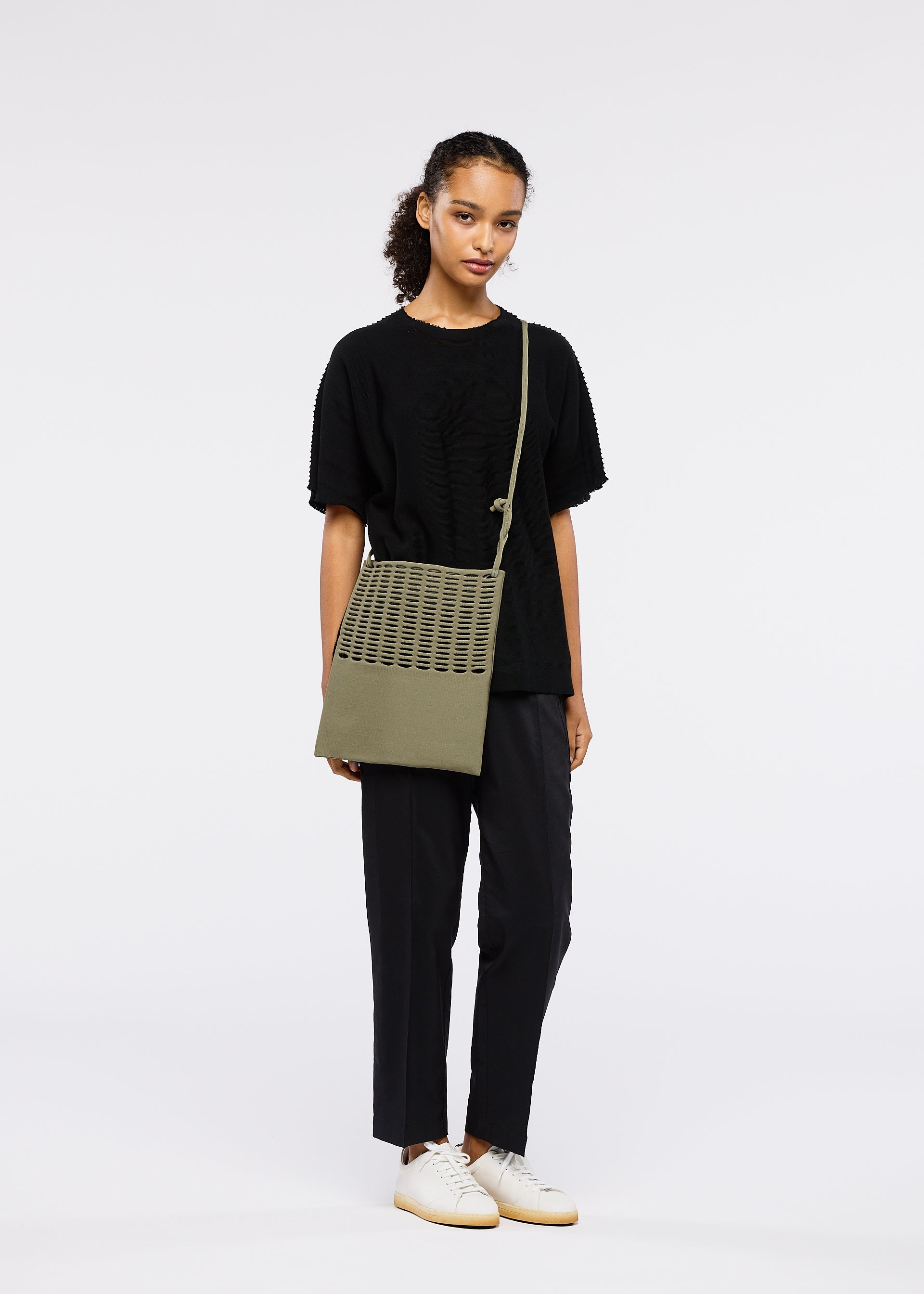 Zoomed view A young woman with curly hair stands against a plain white background, wearing black clothes and white sneakers, holding the olive green MOKKO FLAP POUCH by GOOD GOODS ISSEY MIYAKE with a perforated pattern.