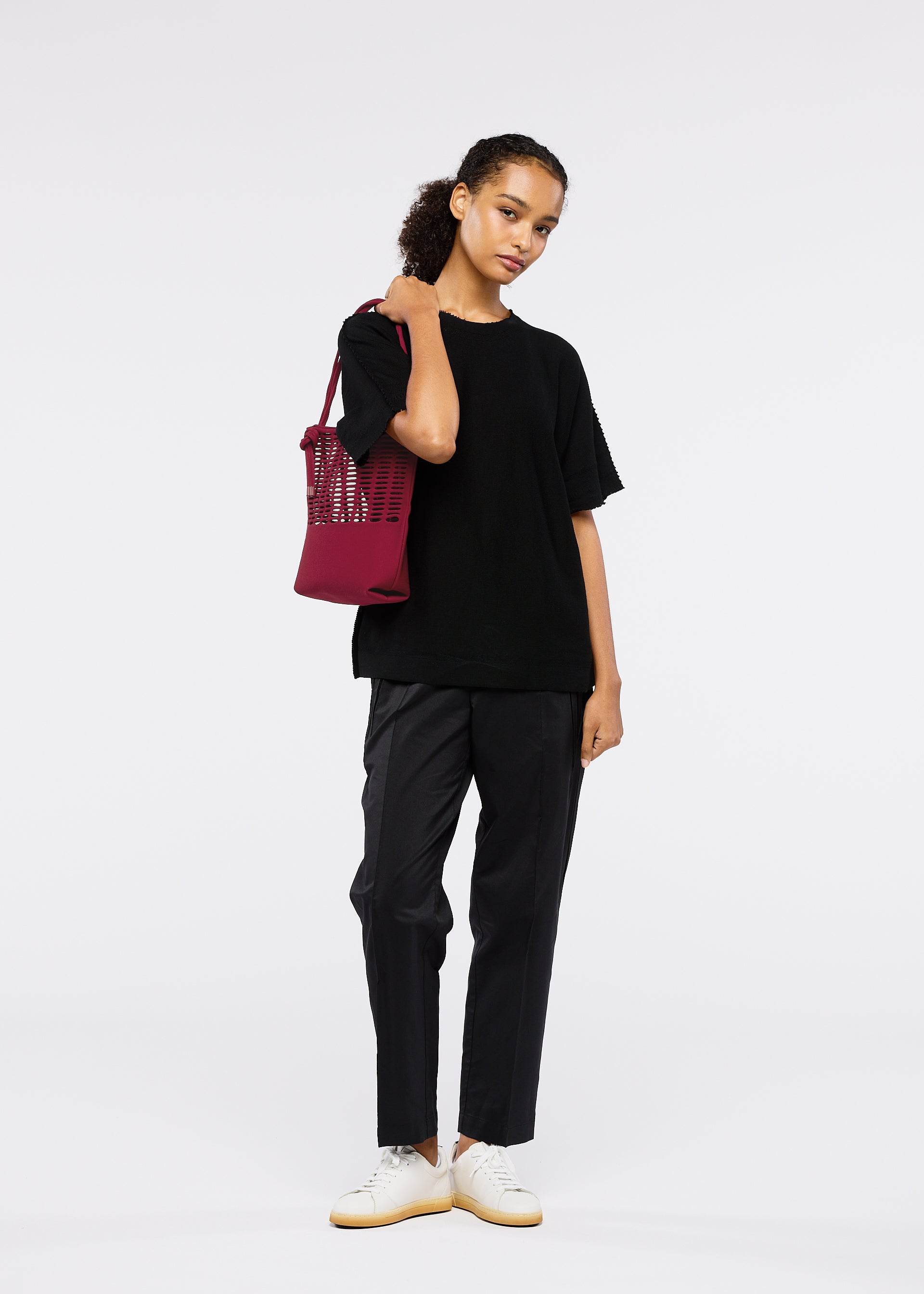 Zoomed view A young person with curly hair stands against a white background, wearing a loose black t-shirt, dark pants, white sneakers, and carrying the GOOD GOODS ISSEY MIYAKE MOKKO FLAP POUCH over one shoulder.