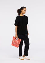 A woman stands against a white background in black T-shirt and pants with white sneakers, holding the coral MOKKO S.C. knit tote bag from the GOOD GOODS ISSEY MIYAKE Spring Summer 2026 collection, looking slightly to the side.
