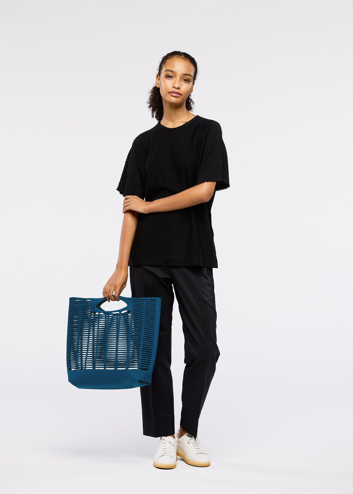 A person stands against a plain background, wearing a black t-shirt, black pants, and white sneakers, holding the MOKKO S.C. tote bag by GOOD GOODS ISSEY MIYAKE—a bold blue washable knit with cutout details, perfect for Spring Summer 2026. view 7 of 8