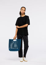 A person stands against a plain background, wearing a black t-shirt, black pants, and white sneakers, holding the MOKKO S.C. tote bag by GOOD GOODS ISSEY MIYAKE—a bold blue washable knit with cutout details, perfect for Spring Summer 2026.