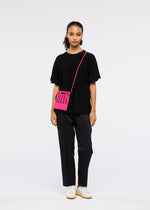 A person faces forward on a plain white background, dressed in a loose black T-shirt, black pants, and white sneakers. They carry the bright pink MOKKO S.C. pochette by GOOD GOODS ISSEY MIYAKE with a detachable strap; their hair is tied back.