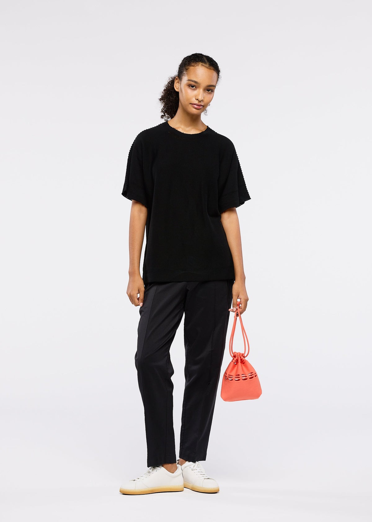 A woman with her hair in a ponytail stands against a plain background, wearing black pants, a loose black T-shirt, white sneakers, and holding the MOKKO S.C. POCKET by GOOD GOODS ISSEY MIYAKE—a trendy, washable knit bag for Spring Summer 2026. view 7 of 7