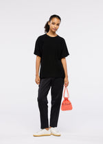 A woman with her hair in a ponytail stands against a plain background, wearing black pants, a loose black T-shirt, white sneakers, and holding the MOKKO S.C. POCKET by GOOD GOODS ISSEY MIYAKE—a trendy, washable knit bag for Spring Summer 2026.