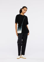 A woman with curly hair pulled back stands against a white background, wearing a black t-shirt, black pants, white sneakers, and the eco-friendly MOKKO NET GRADATION pochette by GOOD GOODS ISSEY MIYAKE. She gazes slightly to the side.