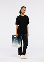 A woman poses against a white background in black attire and white sneakers, holding the MOKKO NET GRADATION tote by GOOD GOODS ISSEY MIYAKE, a modern recycled fishing net bag with cut-out pattern and blue-to-white gradient.