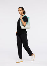 A woman stands against a white background, wearing black clothes and white sneakers. She confidently carries the GOOD GOODS ISSEY MIYAKE MOKKO NET GRADATION tote with a blue-green pattern and looks directly at the camera.