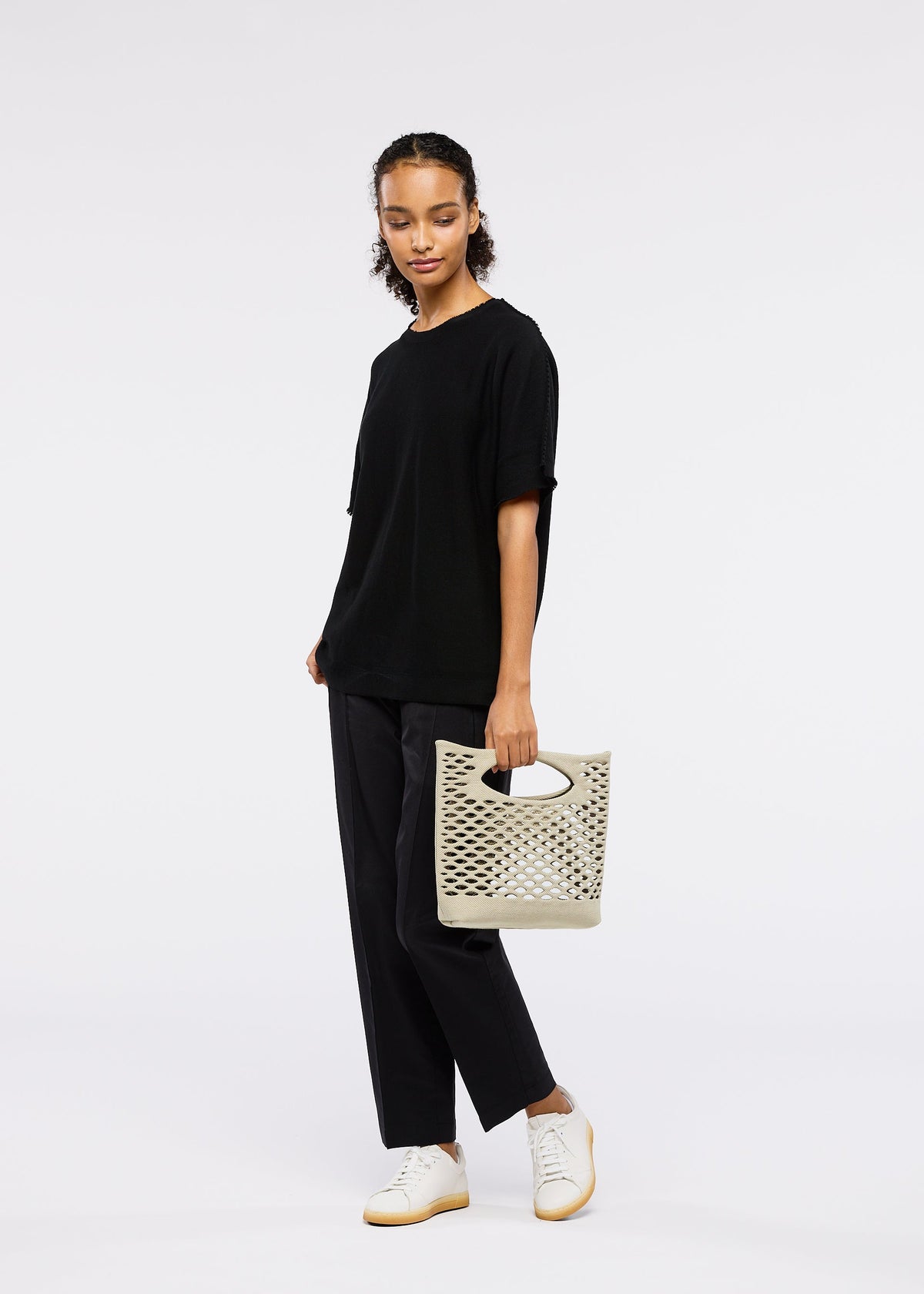 A woman stands against a white background, wearing a black t-shirt, black pants, white sneakers, and holding the MOKKO BASKET tote bag by GOOD GOODS ISSEY MIYAKE. view 5 of 6