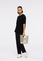 A woman stands against a white background, wearing a black t-shirt, black pants, white sneakers, and holding the MOKKO BASKET tote bag by GOOD GOODS ISSEY MIYAKE.