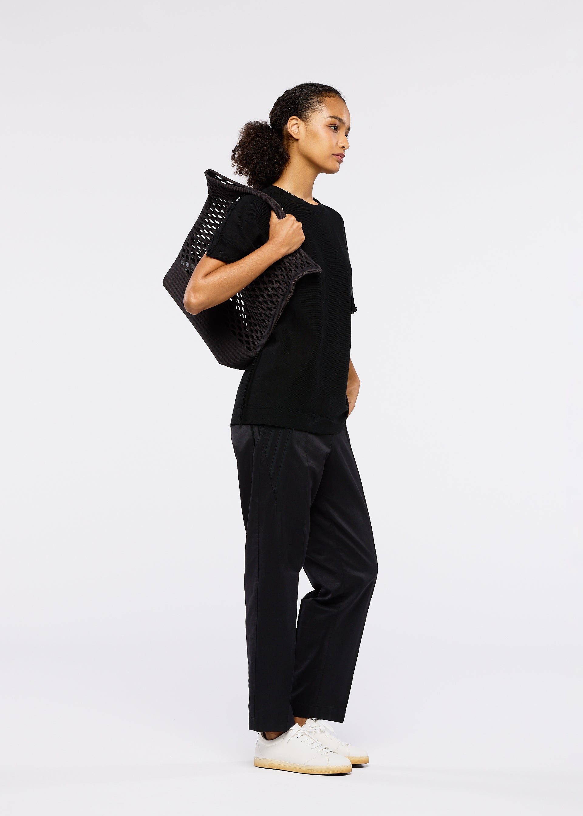 Zoomed view A woman with curly hair tied back stands sideways against a plain white background, wearing a black t-shirt and pants, white sneakers, and carrying the large washable MOKKO BASKET tote by GOOD GOODS ISSEY MIYAKE over her shoulder.
