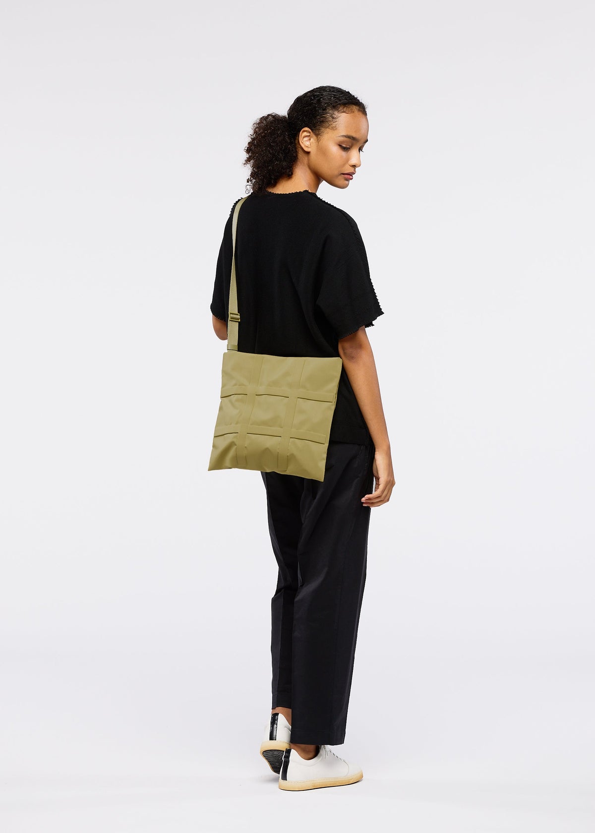 A woman with curly hair stands against a white background, wearing a black outfit and white sneakers, carrying the GRID bag by GOOD GOODS ISSEY MIYAKE in light olive-green over her shoulder. view 9 of 10