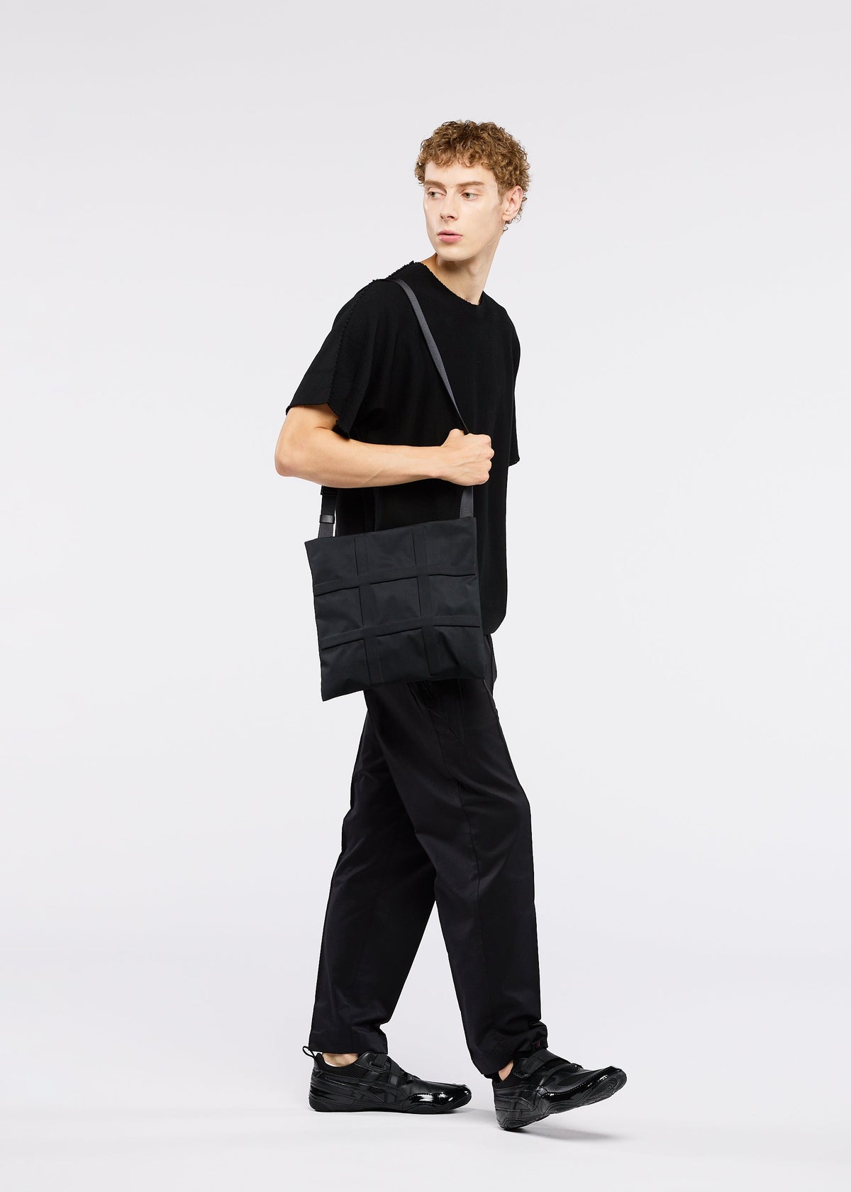 A person with curly hair stands against a plain white background, wearing all black and carrying the GOOD GOODS ISSEY MIYAKE GRID adjustable bag over their shoulder. view 10 of 10