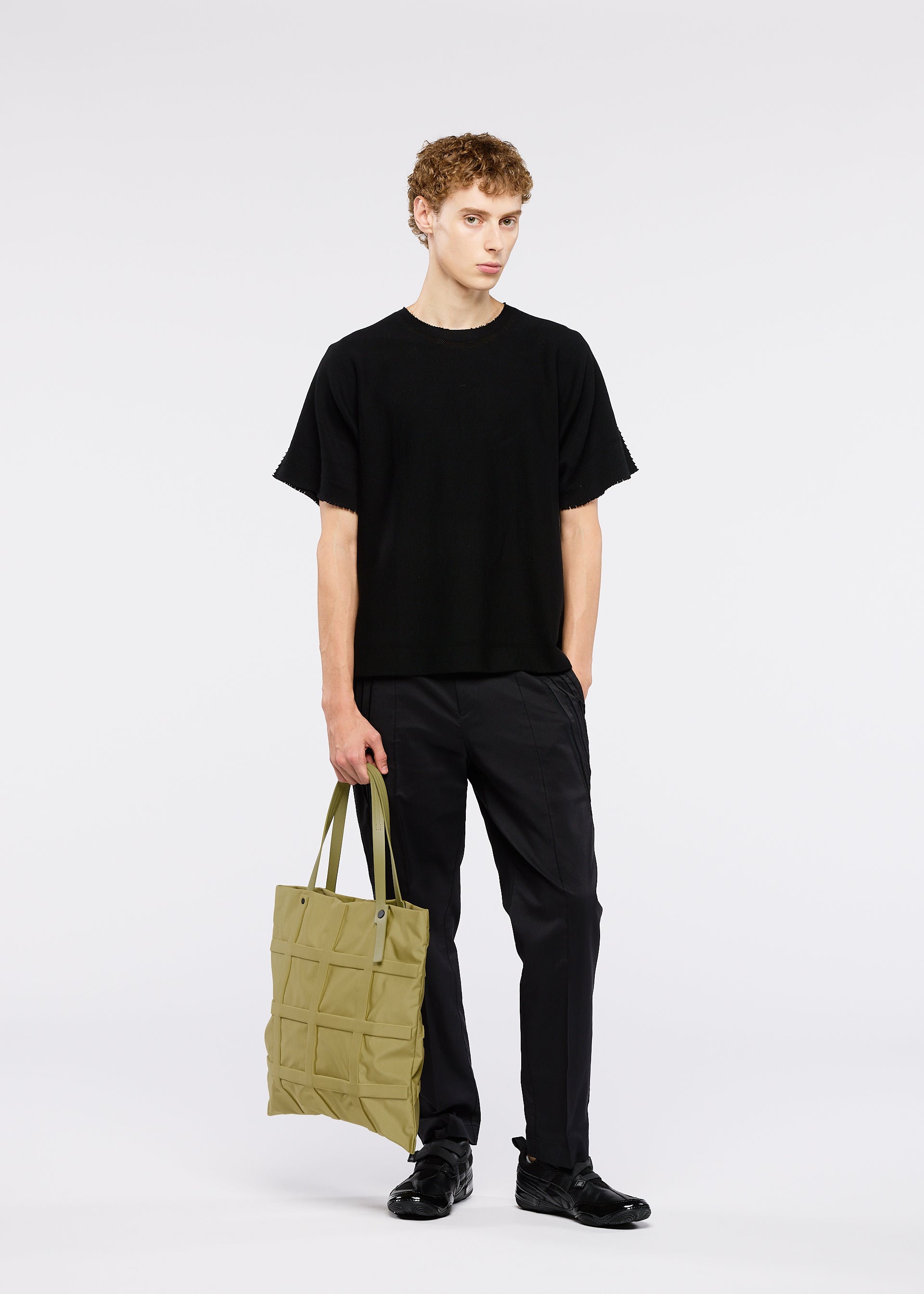 Zoomed view A young man with short curly hair wears a loose black outfit and black shoes, standing against a white background holding the GRID adjustable tote bag in light olive-green by GOOD GOODS ISSEY MIYAKE.