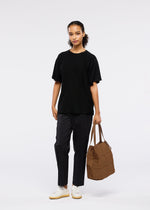 A woman stands against a white background wearing a loose black T-shirt, dark pants, white sneakers, and holds the adjustable GRID tote bag by GOOD GOODS ISSEY MIYAKE in her left hand while calmly looking at the camera.