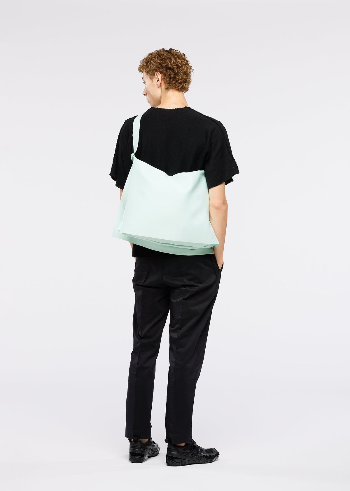 A person with short curly hair dressed in black, facing away with hands in pockets, carries the large pale green ROLL bag by GOOD GOODS ISSEY MIYAKE over their shoulder. The background is plain white. view 6 of 6